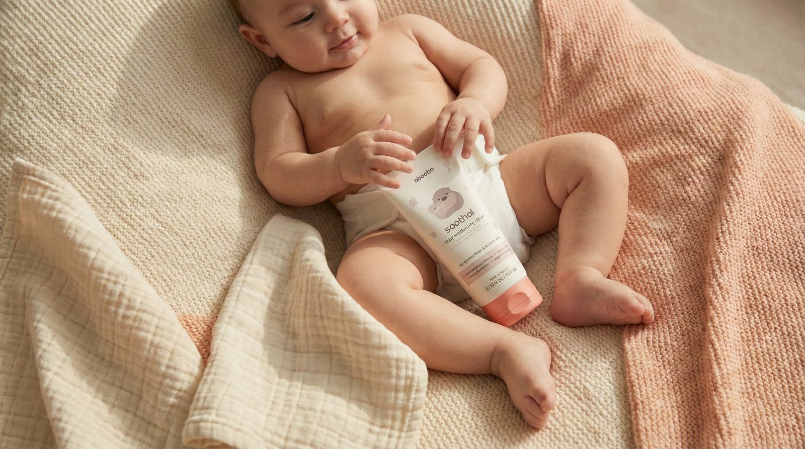 A baby lying on a soft knit blanket holding a tube of SOOTHAL cream